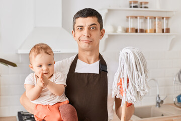 Wall Mural - Indoor shot of Caucasian brunette man and infant baby daughter with floor mops in kitchen, father wearing brown apron, looking at camera with funny facial expression, being tired of cleaning house.