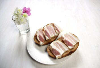 Wall Mural - Salo or salty pork with fat slices on brown bread on plate with vadka glass and flowers on white background