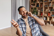 Excited Middle Eastern Man Talking Emotionally On Cellphone, Sitting In Chair At Home, Having Phone Conversation
