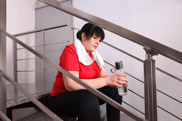 Poster - Tired overweight mature woman with towel and bottle of water sitting on stairs indoors