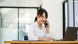 © makibestphoto - Asian Woman entrepreneur busy with her work in the office. Young Asian woman talking over smartphone or cellphone while working on computer at her desk.