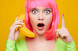 © khmelev - Surprised young sexy woman on pink wig with open mouth call by banana and shows up finger on yellow background