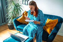 Focused 30s Woman Holding Paper Document Calculating Rent Or Money Savings, Paying Bills In Mobile Application On Cell Phone Doing Monthly Paperwork Sitting On Couch At Home