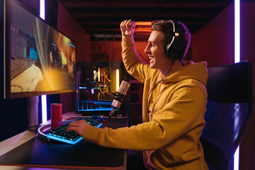 Wall Mural - Young caucasian handsome pro gamer feel excited while playing in online cyber sport game on his powerful PC at home, he making yes hand gesture because he just won in online cyber sport tournament