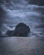 Vertical Shot Of An Ocean Wave Hitting A Huge Rock With A Man Standing Behind It On A Cloudy Day
