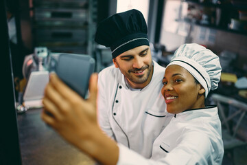 Wall Mural - Happy black chef and her male coworker taking selfie with smart phone at work.