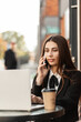 © alones - Happy beautiful hr manager woman in a fancy black suit with a tie and shirt works on a laptop and talks on the phone, sitting and drinking coffee in a cafe in town