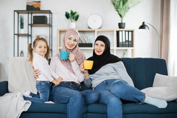 Wall Mural - Three muslim female generations family portrait. Cute girl, islamic mom and grandma hold mugs of hot drinks. Mother, grandmother and granddaughter on couch at home, enjoying leisure time together