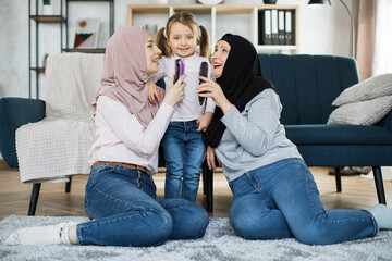 Wall Mural - Three generations of Muslim women sitting on their knees on the floor near the sofa in the living room at home and sing their favorite songs using a comb instead of a microphone.