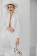 © sergiokat - Confident young blond woman smiling, looking at camera isolated on white background. Studio portrait of successful friendly female in white suit and hat, posing over white wall.