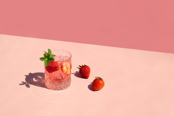 Minimal concept a glass of cocktail with strawberries and ice cubes on pink background. Summer drink and fruits on sunny day.