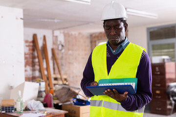 Responsible african american supervisor in yellow safety vest and hard hat inspecting construction site indoors, checking adherence to plans and blueprints on laptop ..