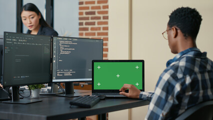 Database designer writing code using laptop with green screen chroma key mockup sitting at desk in busy developers agency. Programer looking at multiple screens with programming language.