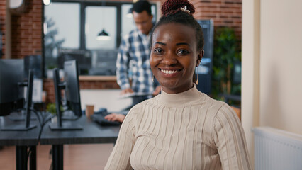 Portrait of african american programer sitting down coding on laptop looking up and smiling at camera. Coder using portable computer next to mixed team in software development agency.