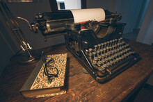 An Old Classic Typewriter In A Vintage Style Studio With A Wooden Desk Lit By A Lamp, Wit Old Books All Around. A Moody, Dark Atmosphere For A Writer Working At Night.