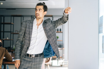 Wall Mural - Portrait of confident elegant business man wearing fashion suit standing by window in modern office room, looking away. Front view of bearded businessman posing at workplace.