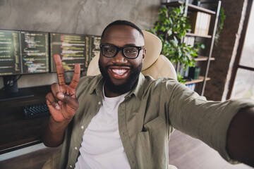 Self-portrait of attractive cheerful funky guy tech company expert showing v-sign at workplace workstation indoors
