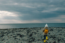 Little Girl In Yellow Rubber Boots Goes To The Sea Along A Rocky Beach In Spring. View From The Back Child With A Yellow Backpack On His Back Walks