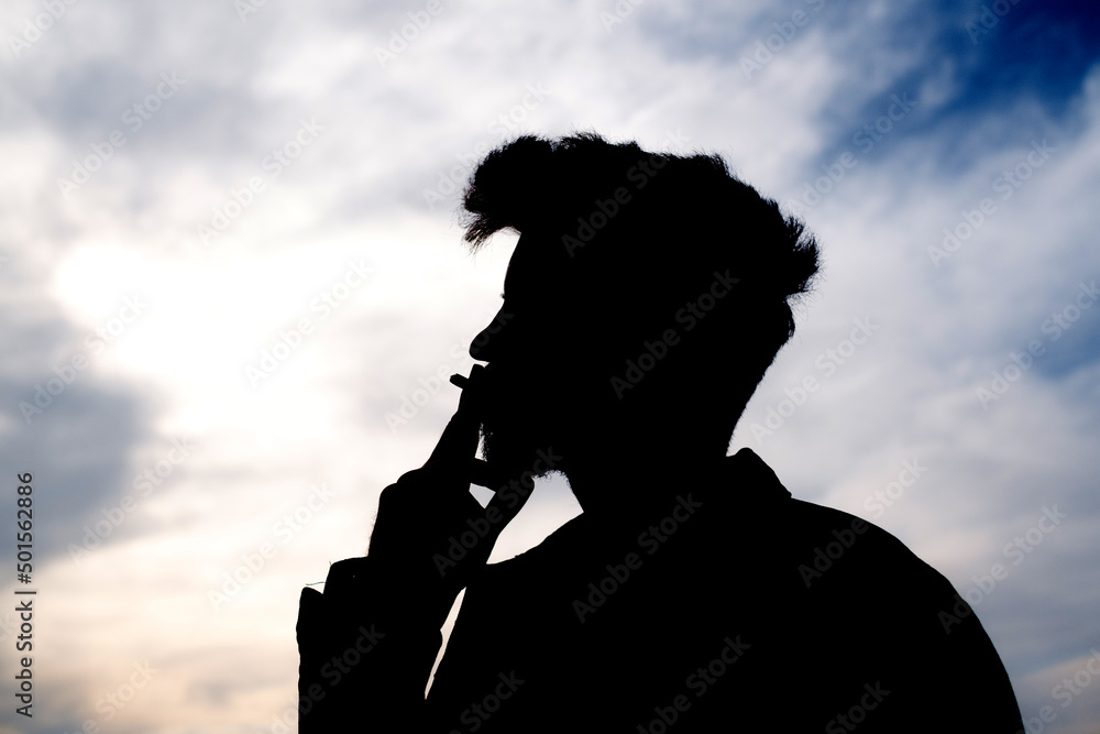 A young man smoking silhouette. One caucasian man portrait smoking  cigarette silhouette on a park bench outdoor. Thinking about life, sad man.  Stock Photo