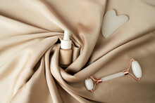 A Face Massage Cream, A White Guasha Face Massager In The Form Of Heart And Face Roller Lying On A Beige Fabric