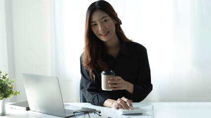 Smiling Asian businesswoman holding a coffee mug and laptop at the office. Looking at the camera.