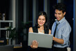 © Stella - Happy smiling young Asian man and woman teammate officer have overwork project overnight in office, use laptop computer for discussing job at mid night, work  hard overload night-shift business people