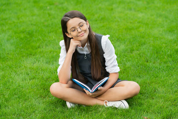 dreamy child in glasses reading book sitting on green grass