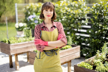 Portrait of young and cheerful gardener in green apron standing at home vegetable garden in backyard. Concept of homegrowing and leisure time