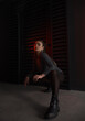 © NASTYA PALEHINA - teen fashion. young brunette girl in a shiny silver bodysuit and boots sits fashion on floor with hands on the knees near black wall background in studio with red light. fashion concept, free space