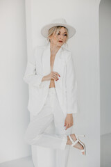 Wall Mural - Confident young blond woman smiling, looking at camera isolated on white background. Studio portrait of successful friendly female in white suit and hat, posing over white wall.