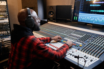 Wall Mural - stylish african american man wearing red checked shirt and headphones working on music track in reco