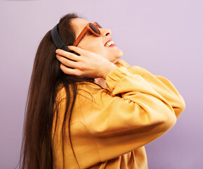 Canvas Print - Fun enjoying pretty young woman listen the music with long brow hair looking up in fashion yellow sport hoodie and sun glasses on purple background.