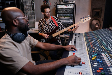 Poster - Young Black musician playing guitar and sound engineer adjusting setting on mixer in recording studio