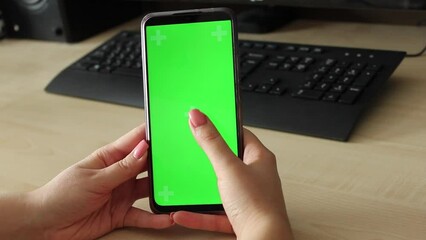 Woman using mobile smart phone with green screen on table with computer and keyboard