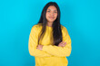 Self confident serious calm young latin woman wearing yellow sweater over blue background stands with arms folded. Shows professional vibe stands in assertive pose.
