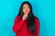 Thoughtful young latin woman wearing red shirt over blue background holds chin and looks away pensively makes up great plan