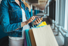Cropped Image Of A Young Caucasian Woman Writing A Message Or Checking Prices In Stores On Her Mobile Phone While Standing With Bags In The Parking Lot