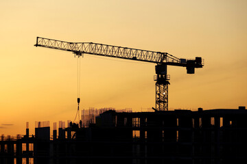 Wall Mural - Silhouette of tower crane with cargo and workers on building scaffolding at sunset. Housing construction, apartment block in city on dramatic sky background