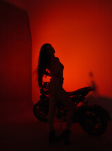 Young Teen Brunette Girl Biker Stands Fashion In Profile Like Silhouette Near Black Sports Motorcycle On A Red Studio Wall Background In Backlight. Teen Leisure. Lifestyle Fashion Concept, Free Space