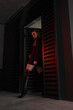 © NASTYA PALEHINA - teen fashion. young girl in black slim dress-jacket and over the knee boots stands fashion in open frame  door at parking on a black metal wall background with red light. lifestyle concept, free space