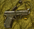 Obrazki ze strzelnicy , pistolet , łuski i zatyczki do uszu ( ochronniki słuchu )  leżące na materiale w kamuflażu 