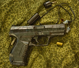 Fototapeta  - Obrazki ze strzelnicy , pistolet , łuski i zatyczki do uszu ( ochronniki słuchu )  leżące na materiale w kamuflażu 