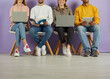 © Studio Romantic - Young people use laptops and notebooks to prepare for lectures, exams and interviews. Legs of multiracial young students sitting in row on chairs on purple wall background. Low section.