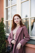 © LIGHTFIELD STUDIOS - redhead young woman in coat standing near windows outside.