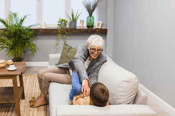 Wall Mural - Grandma and grandson spend beautiful time together, they are happy and enjoy themselves. They tickle on the sofa in the modern living room. Happy grandmother with her grandson in the house.