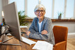 © Dragana Gordic - An attractive positive talented mature woman sits in front of a computer and watches the bills she has to pay, looking at the camera with a happy smile. The senior makes notes in a notebook