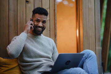 Happy young man with laptop and smartphone resting outdoors in a tree house, weekend away and remote office concept