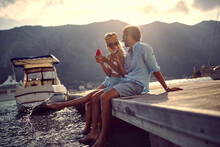 Couple Having Small Talk And Enjoying Watermelon By Water, Sitting On Wooden Jetty. Woman Splashing Water With Her Leg. Boats In Background. Love, Holiday, Lifestyle Concept.