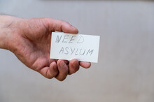Need Asylum. Words Written In Jagged Letters. A Man's Hand Holds A White Paper Rectangle With Text Against A Gray Background.
