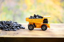 A Small Yellow Toy Truck Is Loaded With Sunflower Seeds Next To A Small Pile Of Sunflower Seeds. A Car On A Wooden Surface Against A Background Of Autumn Forest. Transportation Of Sunflower Seeds